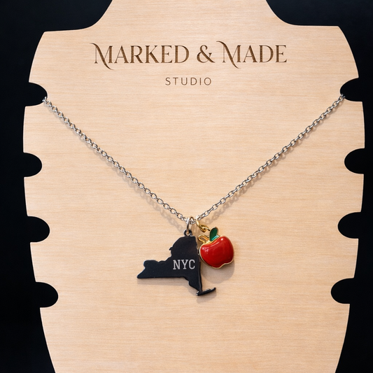 Necklace with a pendant shaped like New York City and an apple on a 'Marked & Made Studio' stand.