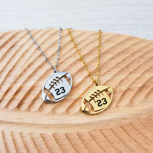 Two football-shaped necklaces, one silver and one gold, with number 23 on a wooden surface.